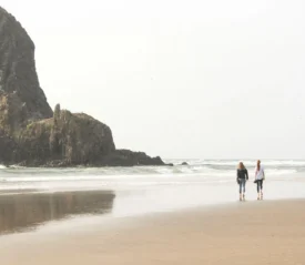 Two people stroll along a wide beach with waves rolling in and a large rocky formation rising from the shoreline nearby