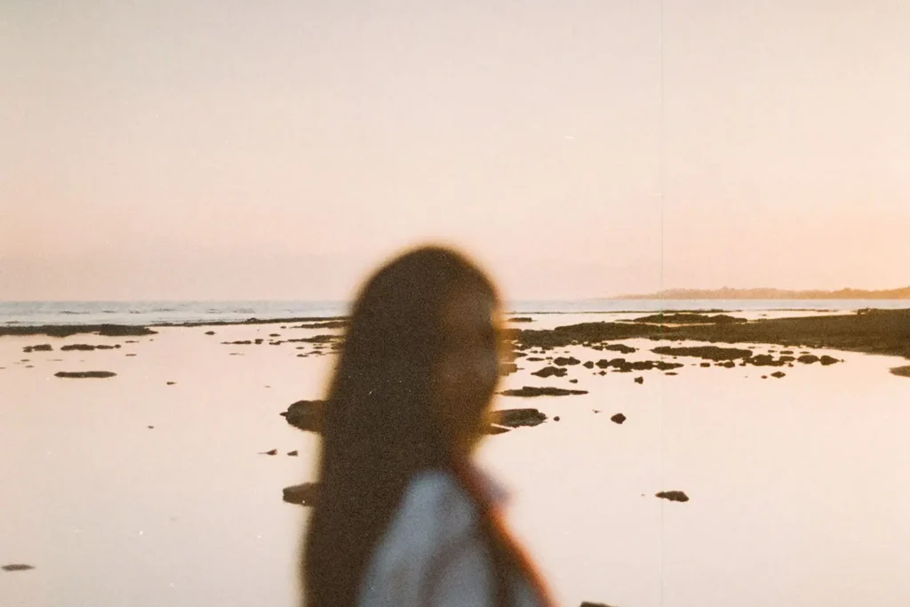 A person stands near the shoreline at sunset, with calm water and scattered rocks stretching into the distance