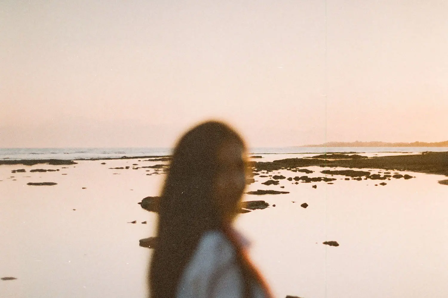 A person stands near the shoreline at sunset, with calm water and scattered rocks stretching into the distance
