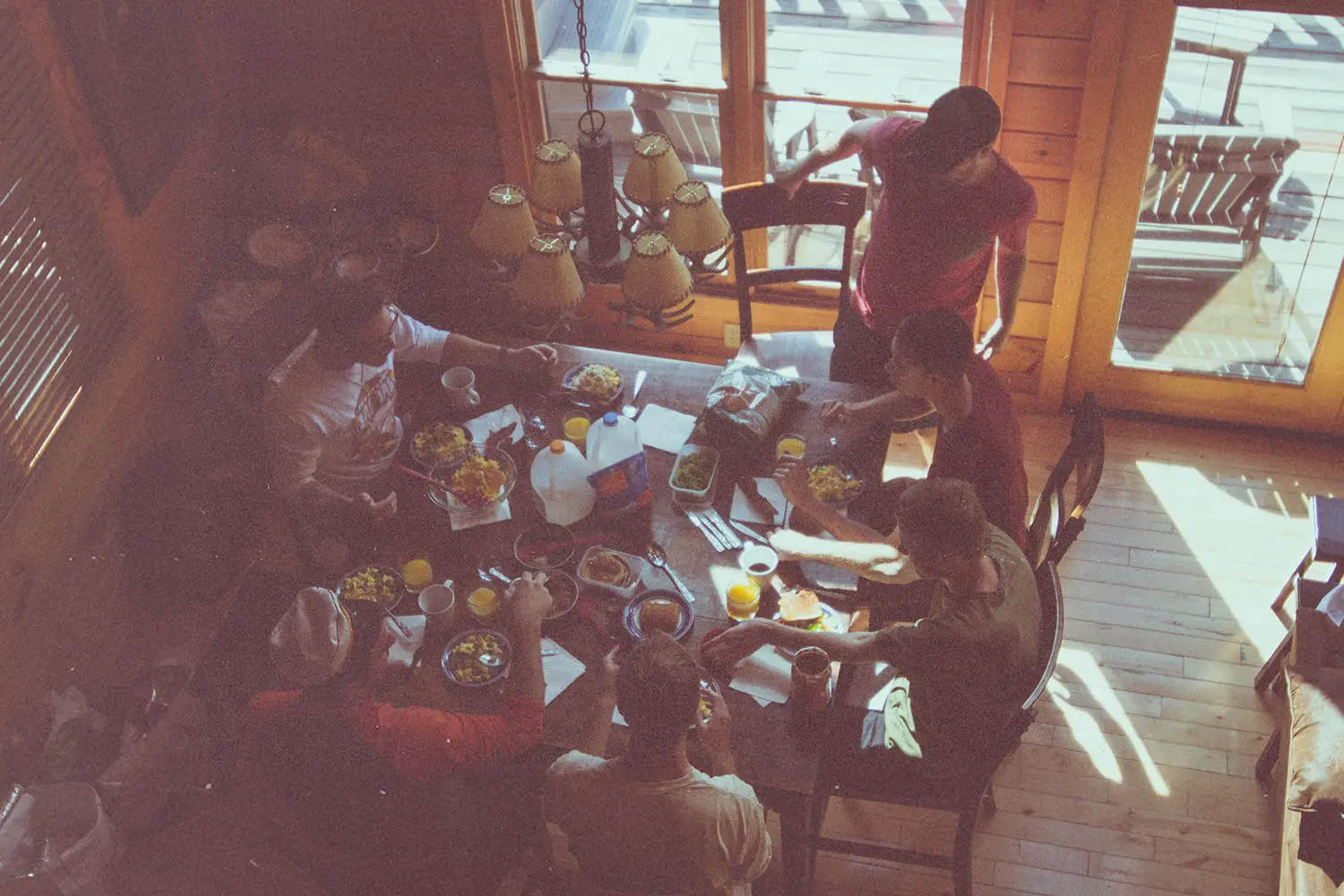 Overhead view of a group gathered around a wooden table sharing breakfast plates, drinks, and snacks inside a sunlit cabin