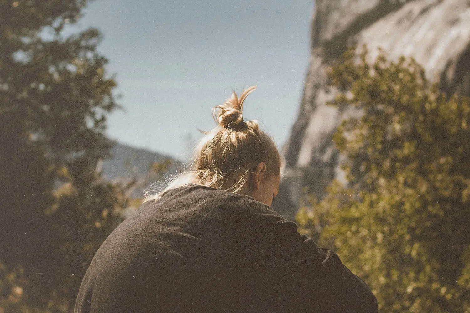 Person sitting outdoors near mountains and trees, viewed from behind on a sunny day