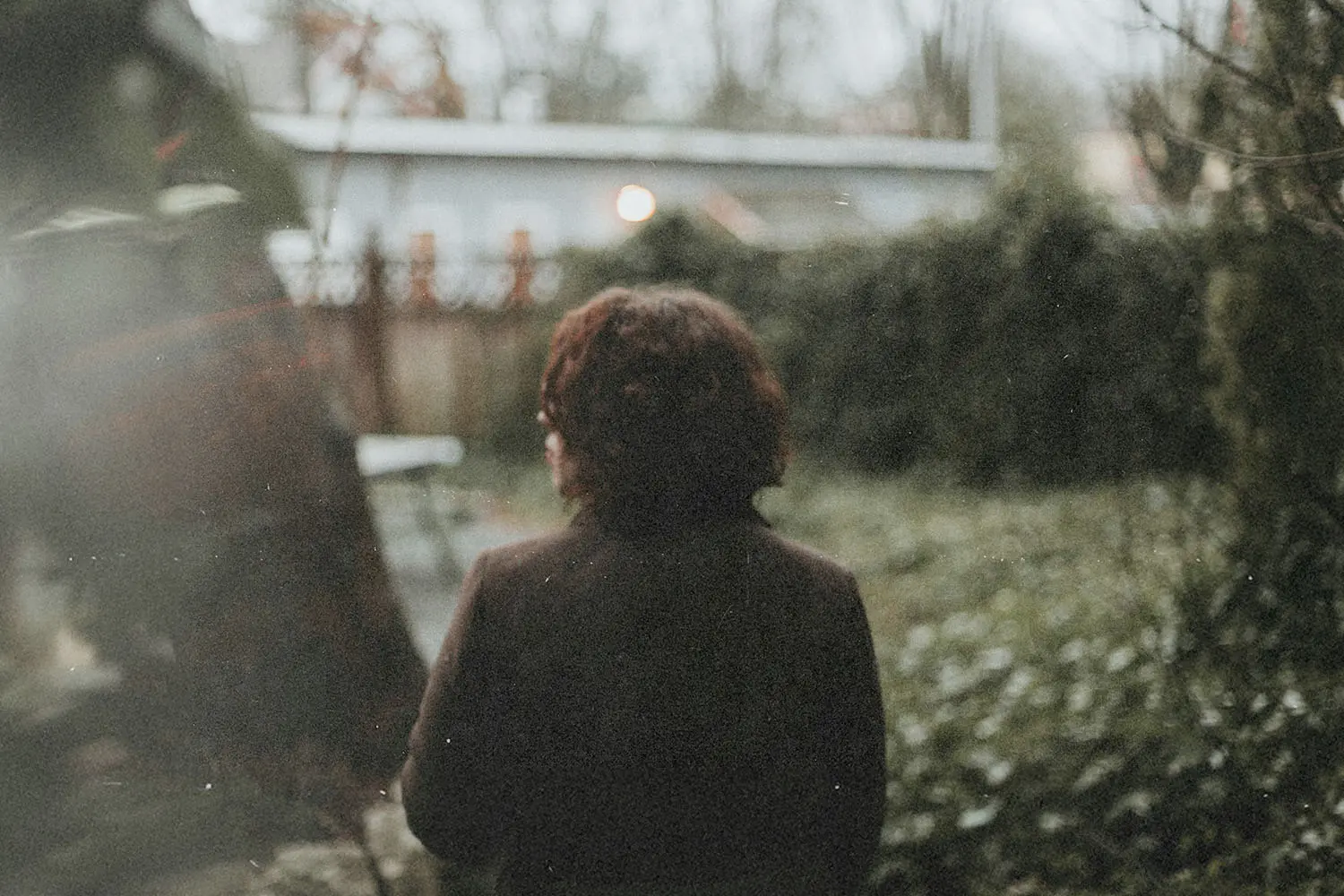 Person standing in a garden facing away from the camera, surrounded by trees and shrubs