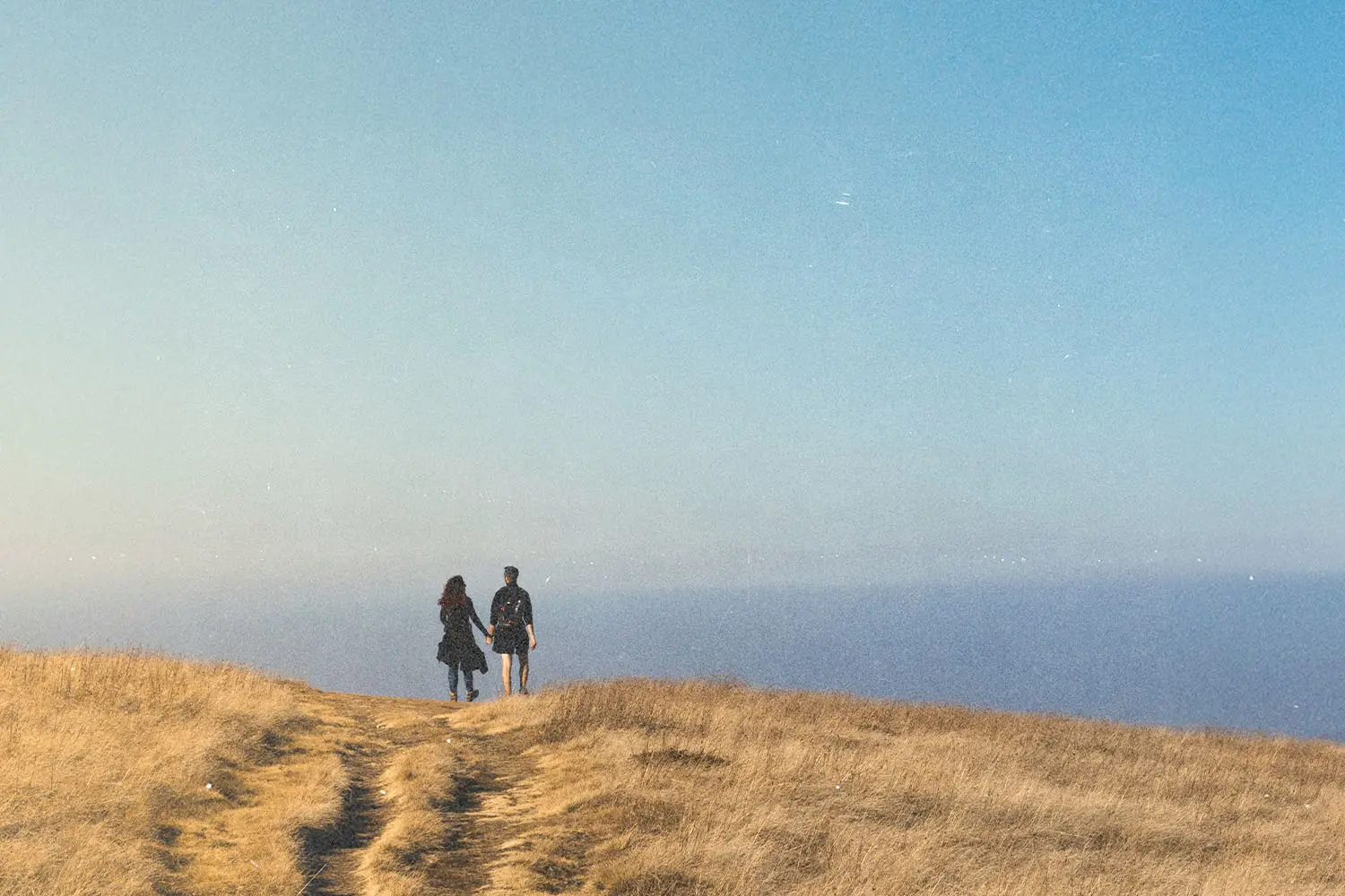 Two people walking along a grassy hilltop trail toward an open horizon beneath a clear blue sky