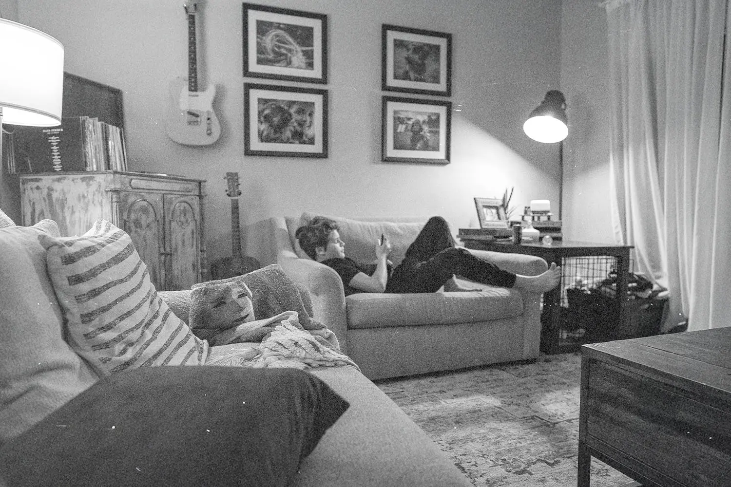 Person lounging on a couch in a cozy living room with framed artwork, a guitar, and soft lighting