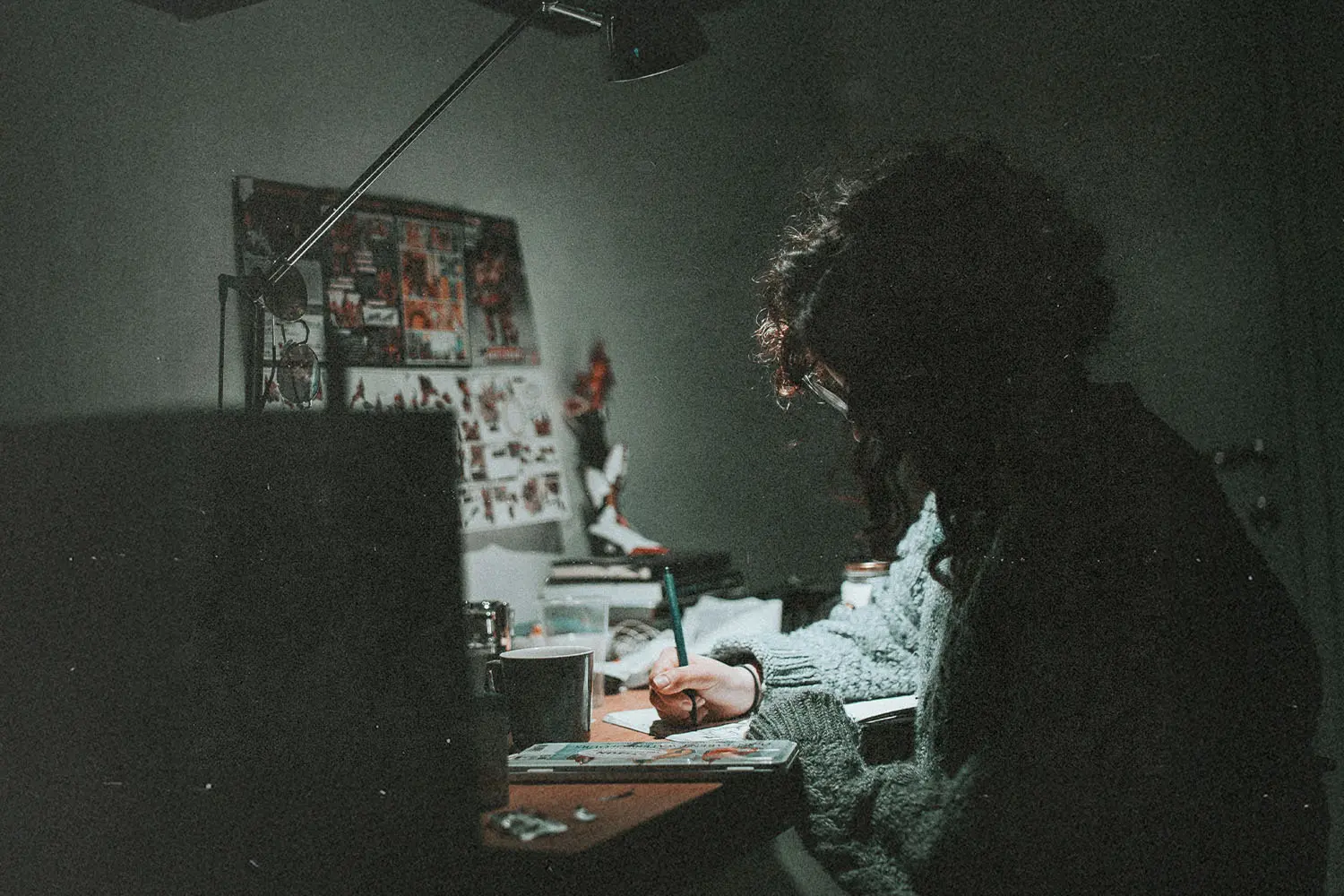 Person seated at a cluttered desk writing under a focused desk lamp in a dimly lit room