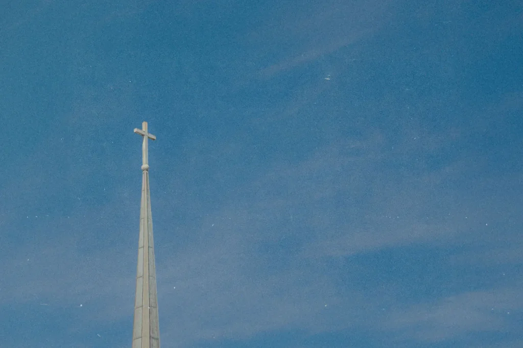 A church steeple with a cross at the top rises into a clear blue sky