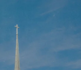 A church steeple with a cross at the top rises into a clear blue sky