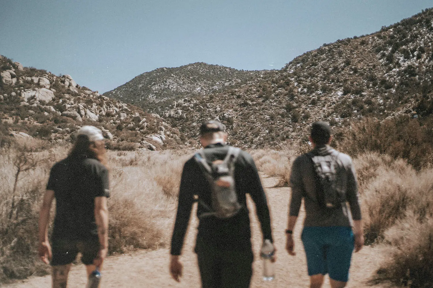 Three hikers walking along a dusty trail toward rugged mountain terrain