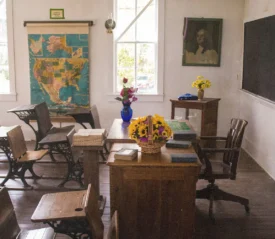 Vintage-style classroom with wooden desks, a teacher’s desk decorated with flowers, a large map on the wall, and sunlight streaming through tall windows