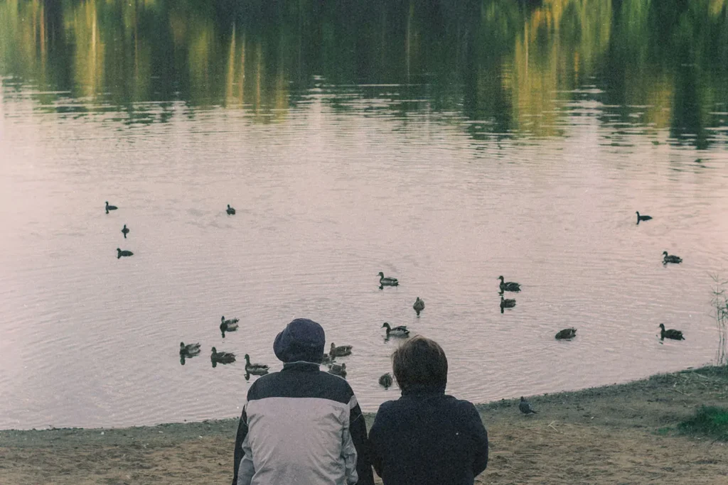 two people sitting at edge of pond watching ducks