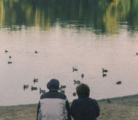 two people sitting at edge of pond watching ducks
