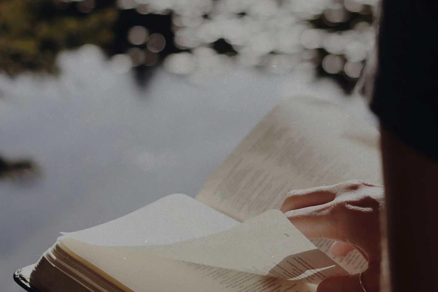 Person holding an open Bible near a body of water with sunlight reflecting off the surface