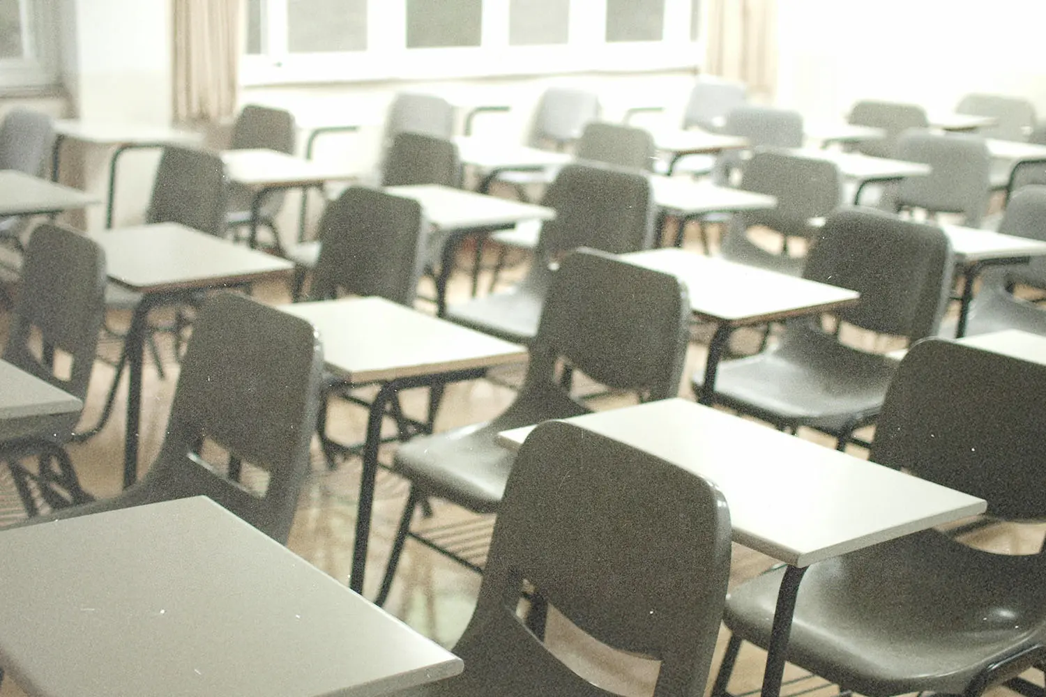 Rows of empty gray student desks in a brightly lit classroom with large windows