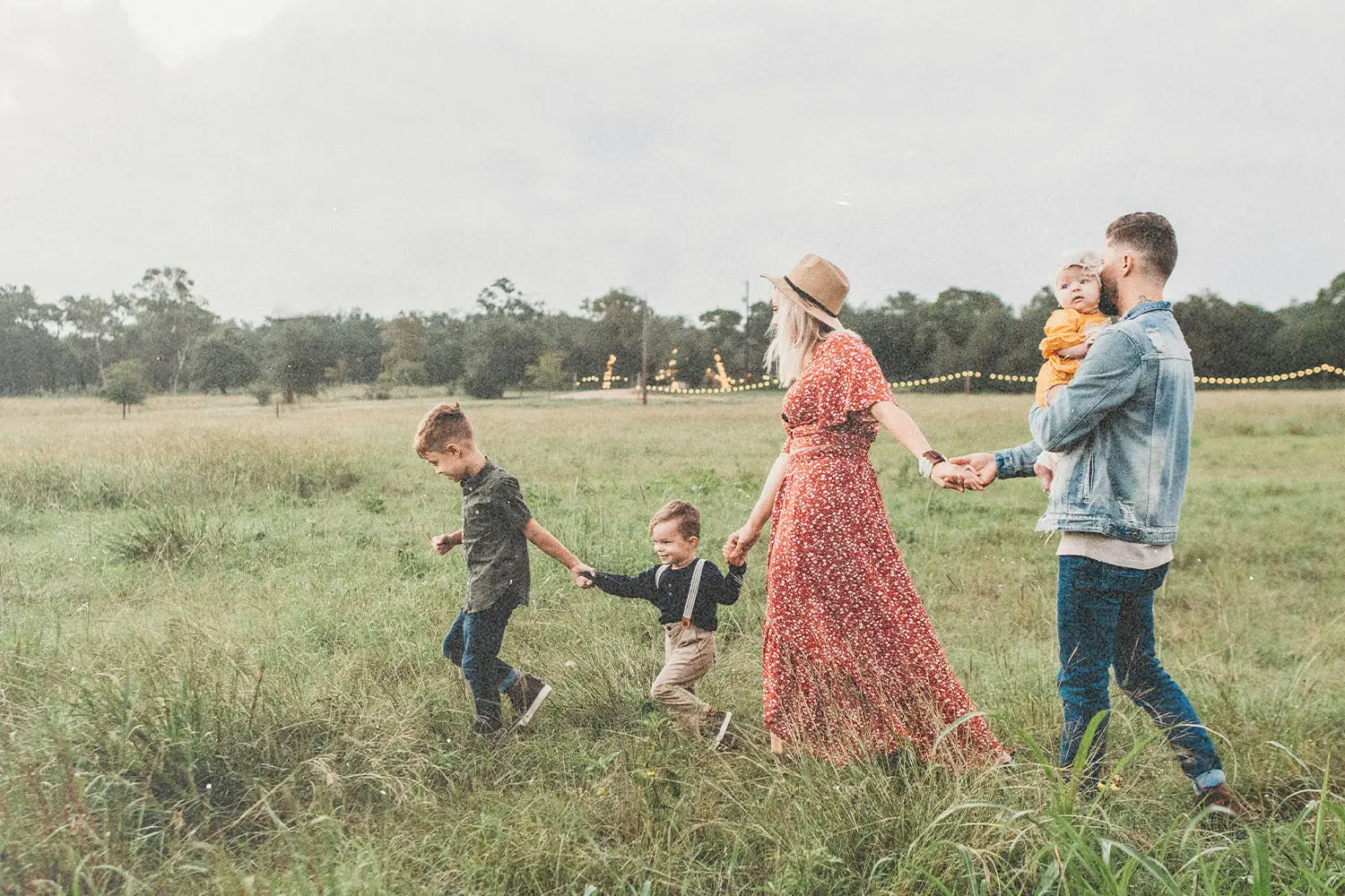 A family walking hand in hand across a grassy field with string lights in the background