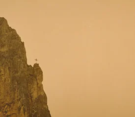A tall rocky cliff rises against a hazy golden sky, with a small cross standing at the edge of the peak