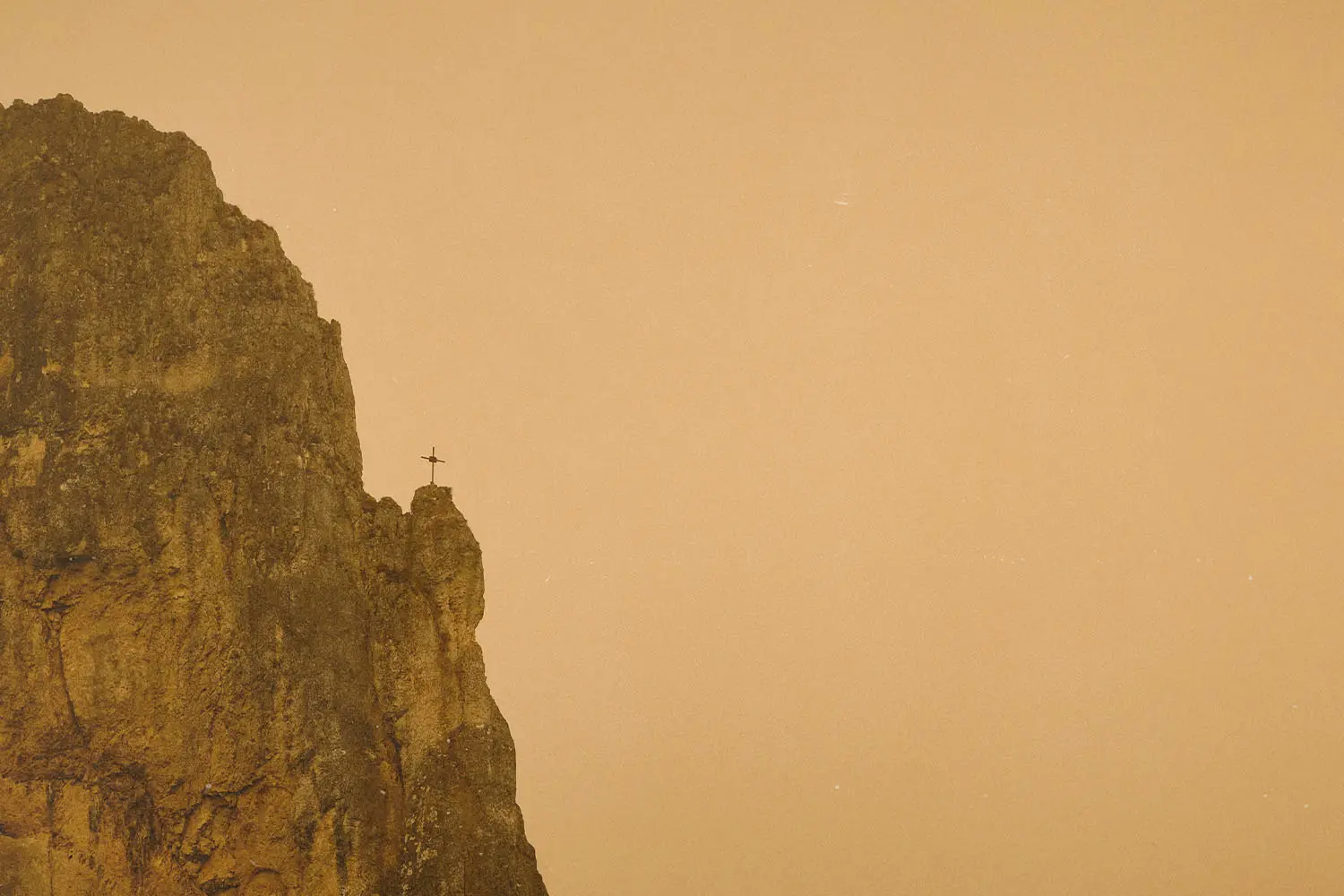 A tall rocky cliff rises against a hazy golden sky, with a small cross standing at the edge of the peak