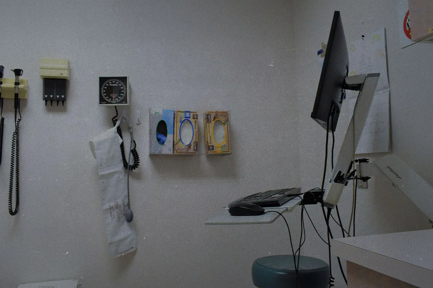 Medical exam room with wall-mounted diagnostic tools, glove boxes, and a computer workstation