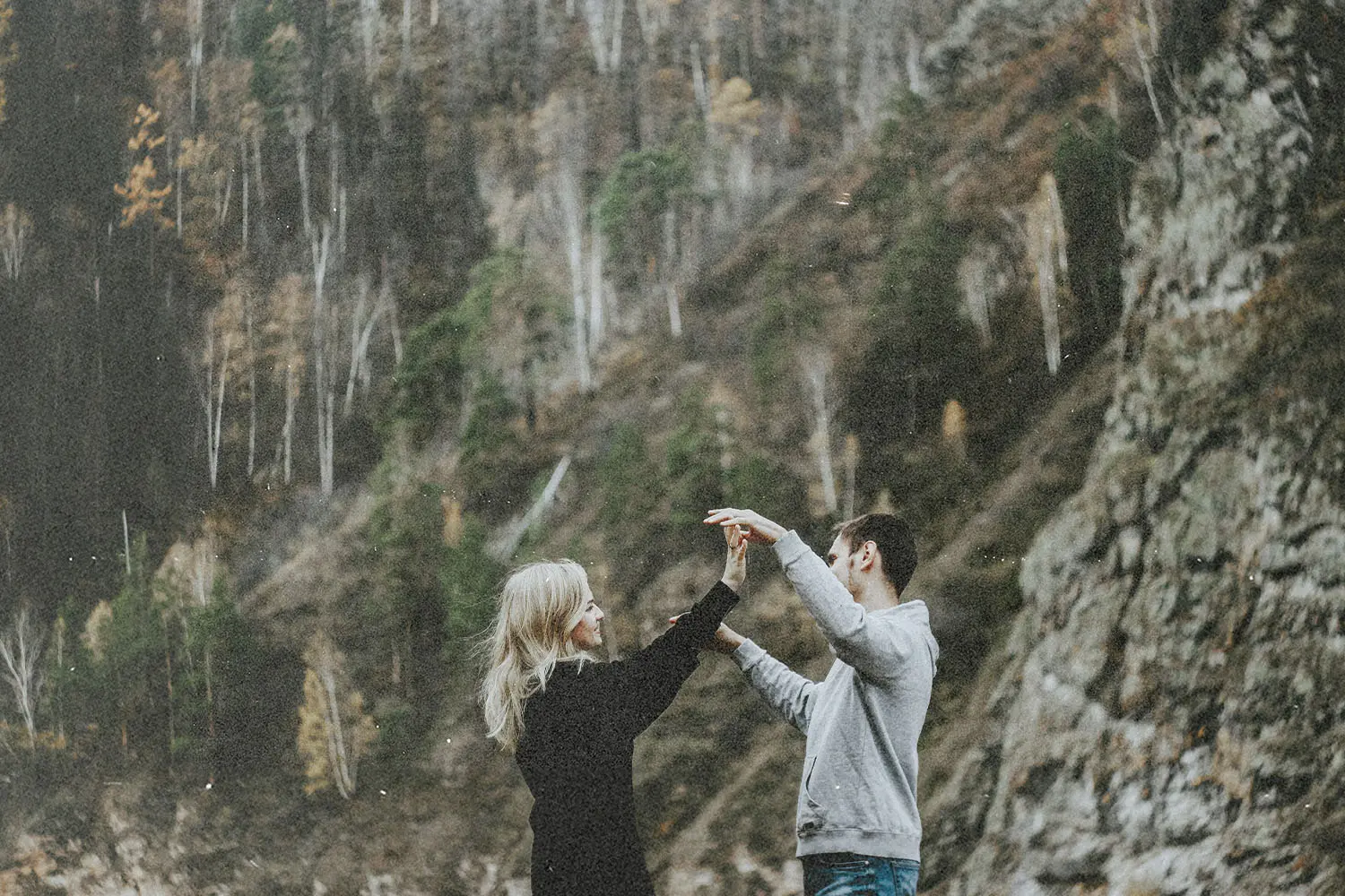 Two people holding hands and standing together in a rocky outdoor landscape with tall trees in the background