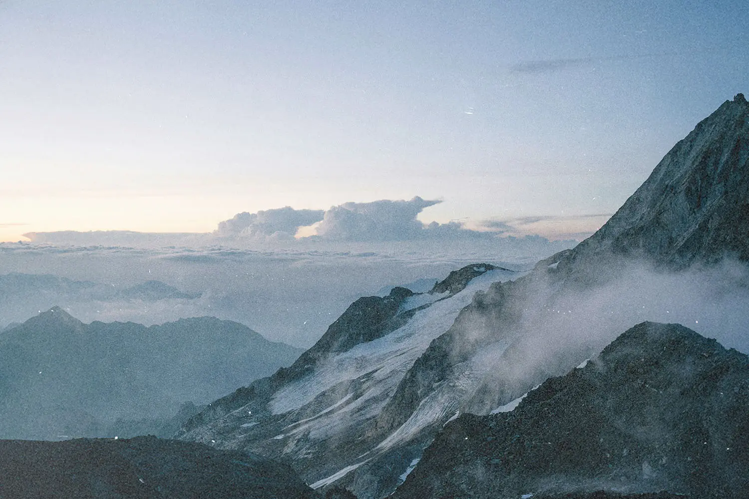 High-altitude mountain range with snow-covered slopes and soft morning light illuminating the clouds.