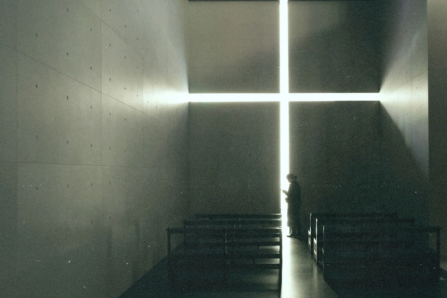 Silhouetted figure standing inside a dimly lit chapel before a large glowing cross-shaped window