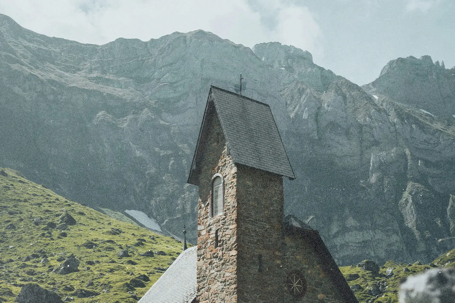 Small stone church with a steep roof set against towering rocky mountains and green hills