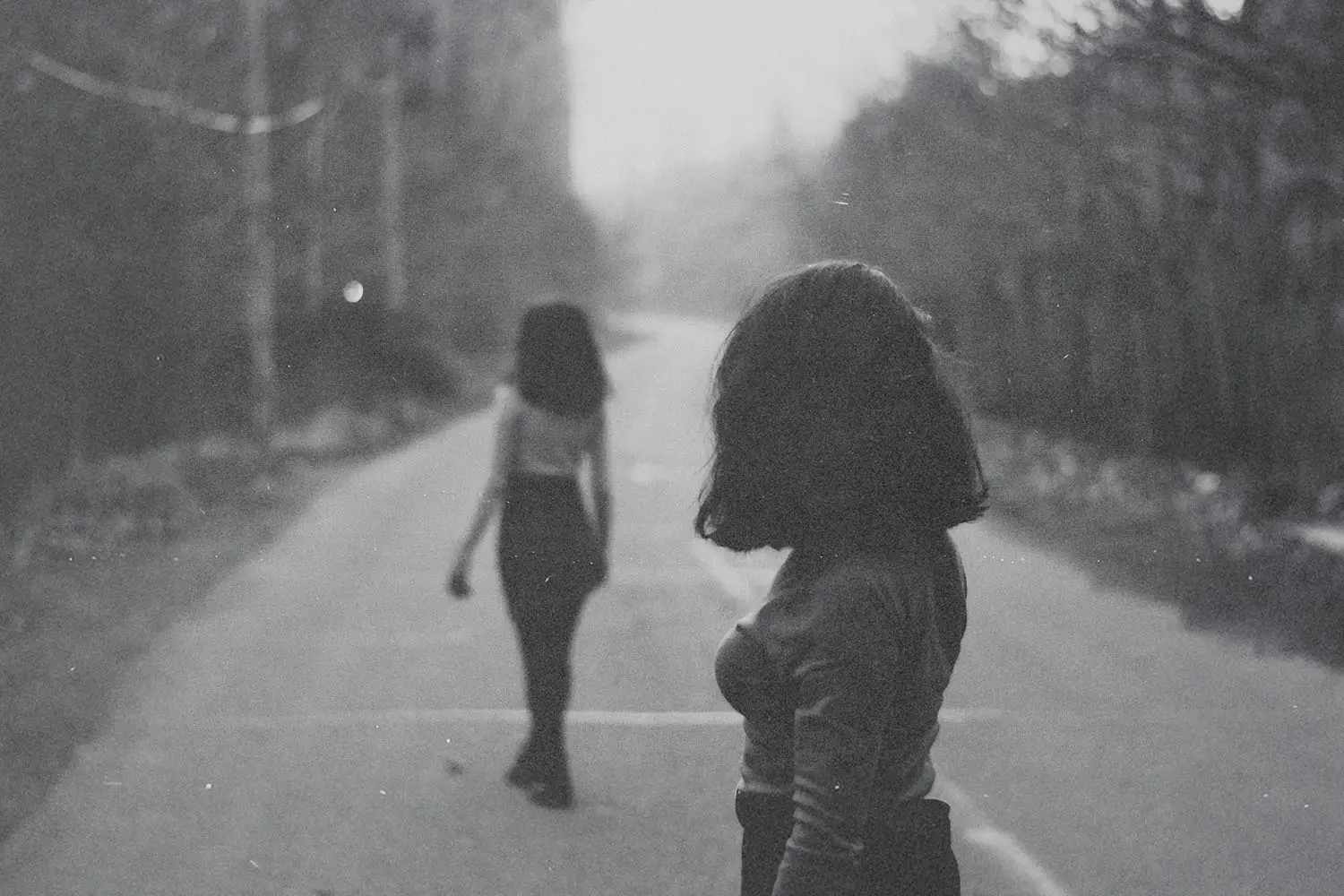 Two people walking on a quiet road lined with trees, one closer to the camera and one farther ahead