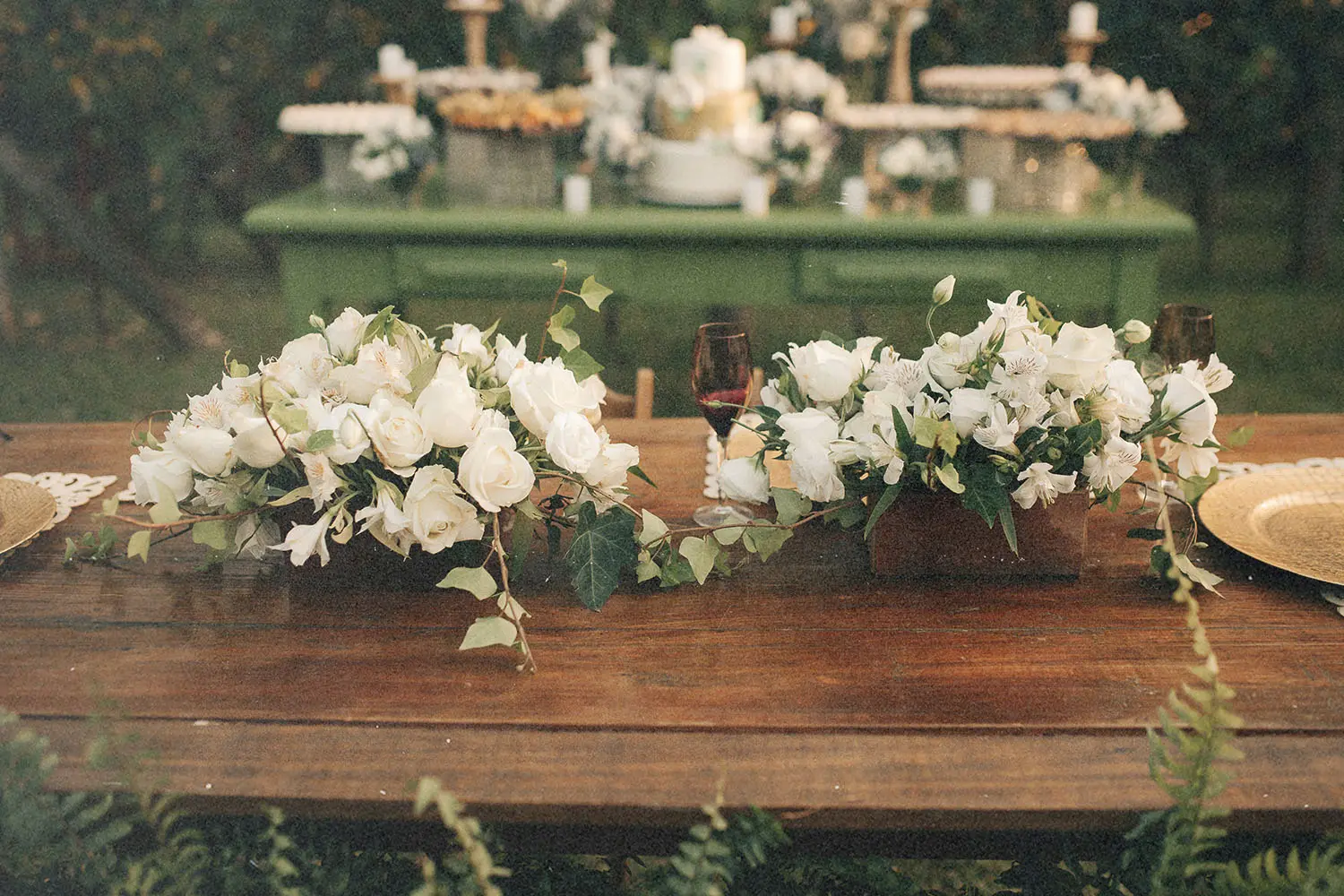 Outdoor wooden table decorated with white floral centerpieces and arranged place settings, with a dessert table in the background