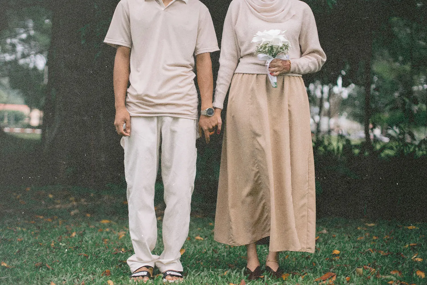 Two people standing outdoors holding hands, with one holding a small bouquet of white flowers