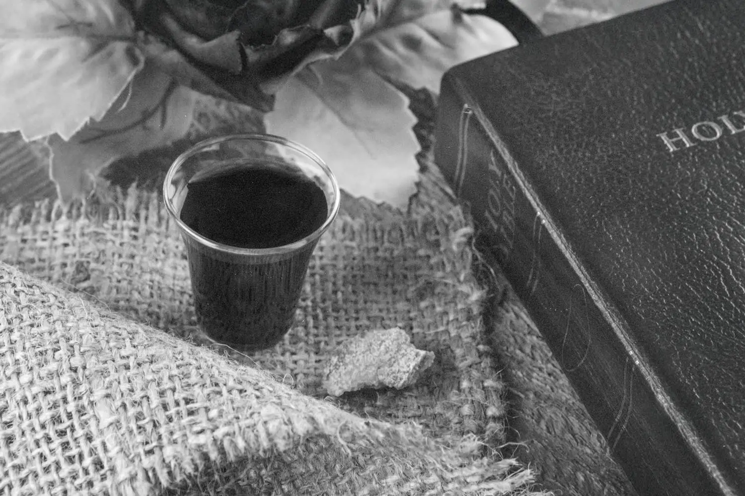 A small cup of communion juice sits on a piece of burlap alongside a broken piece of bread, with a Bible and decorative leaves arranged nearby in a softly lit setting