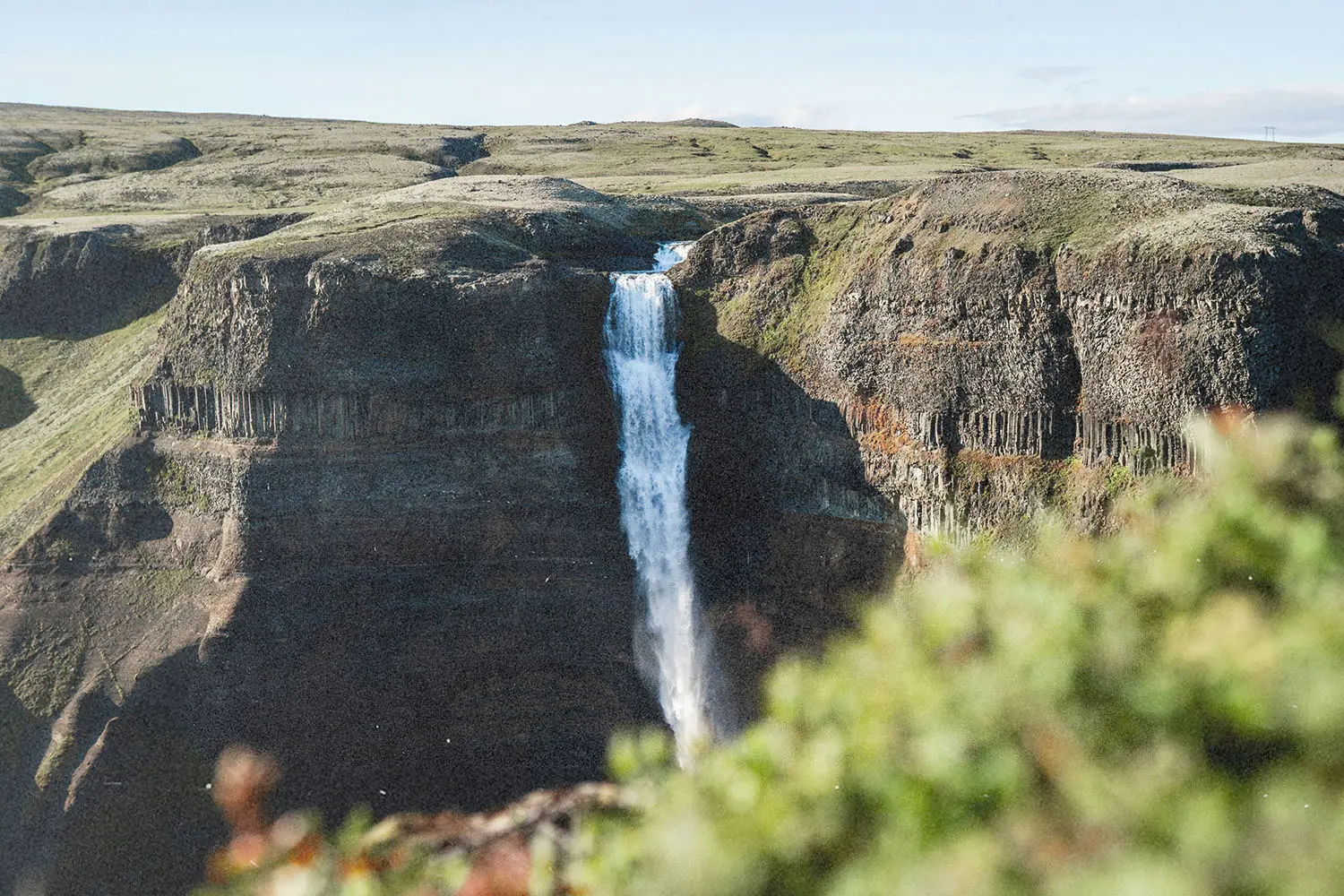A tall waterfall cascades over a steep cliff into a rocky valley surrounded by green terrain
