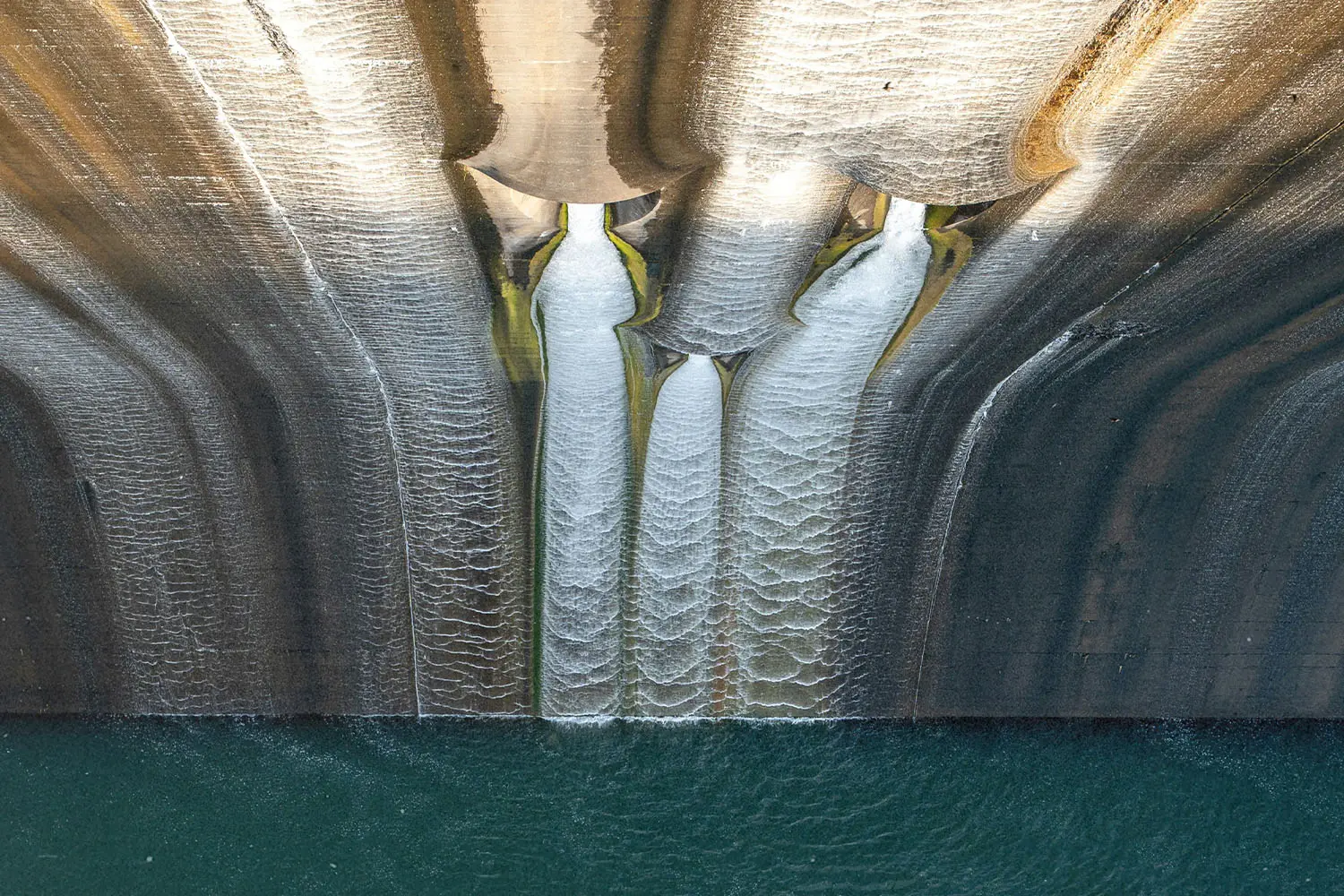 looking down from the top of a dam, water falling down a concrete wall into body of water