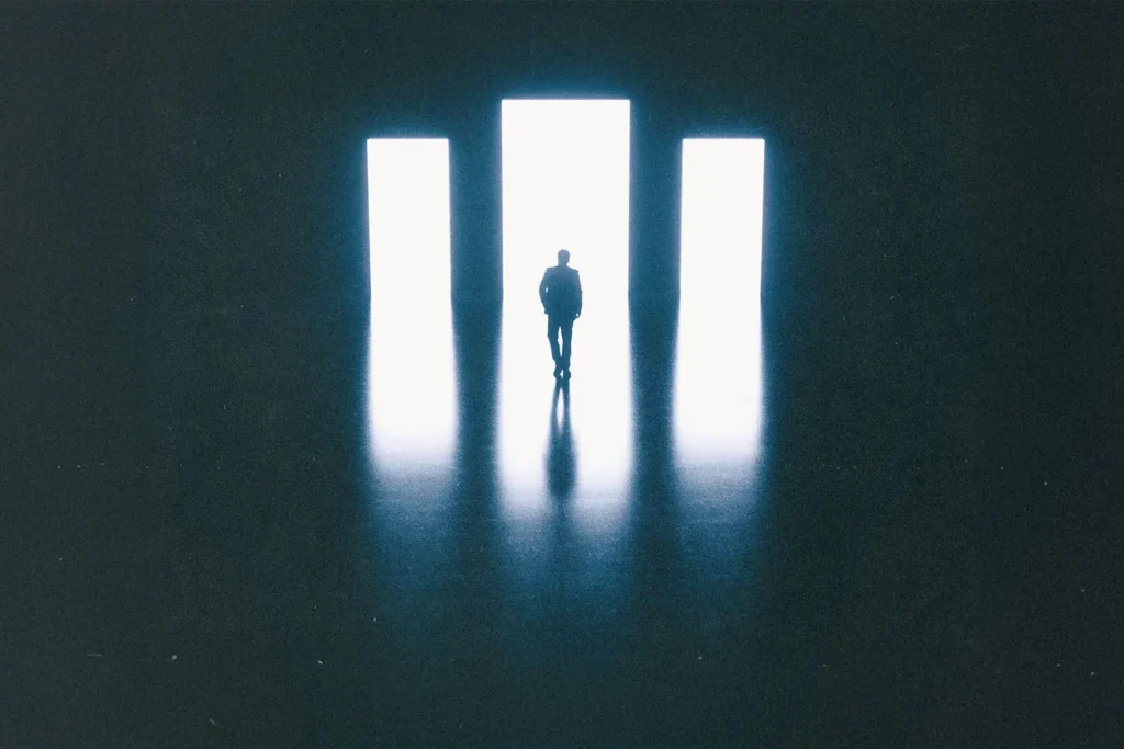 Silhouette of a person standing before three tall, bright rectangular doorways in a dark room, with light reflecting across the floor