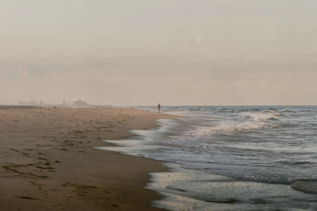 A lone person walks along a quiet shoreline as gentle waves roll onto the sand in the early morning light