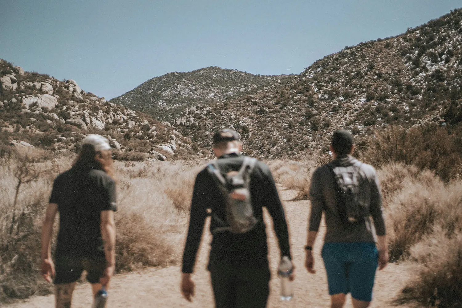 Three people walk along a dirt trail surrounded by dry brush, with rocky mountains rising in the background under a clear sky