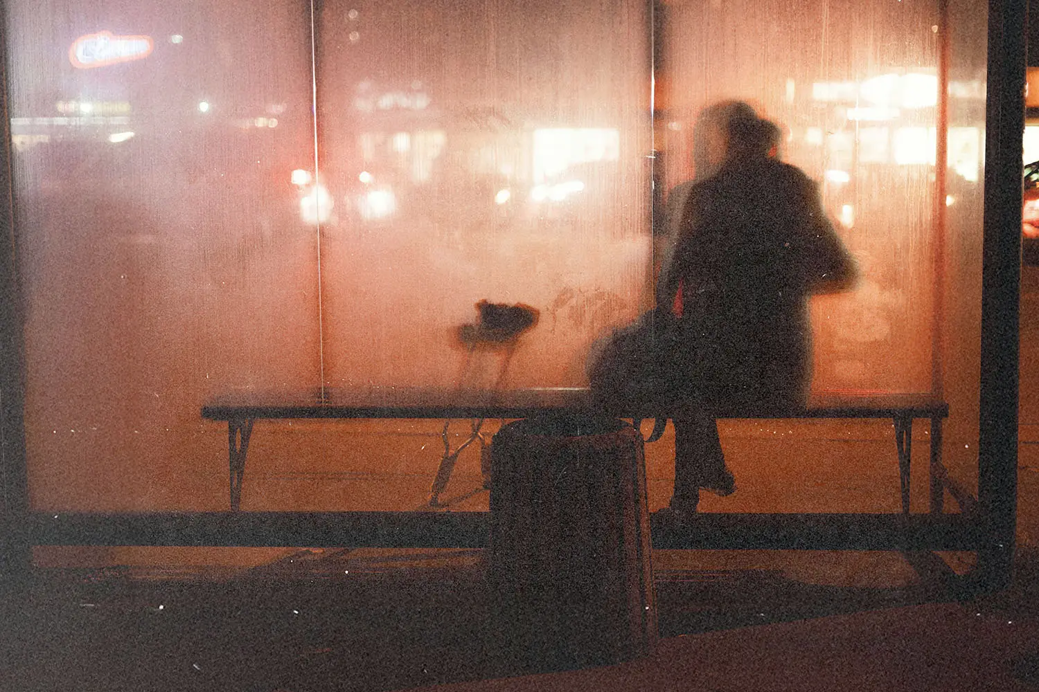 A person sitting on a bench behind a fogged glass wall at night with city lights visible through the window.