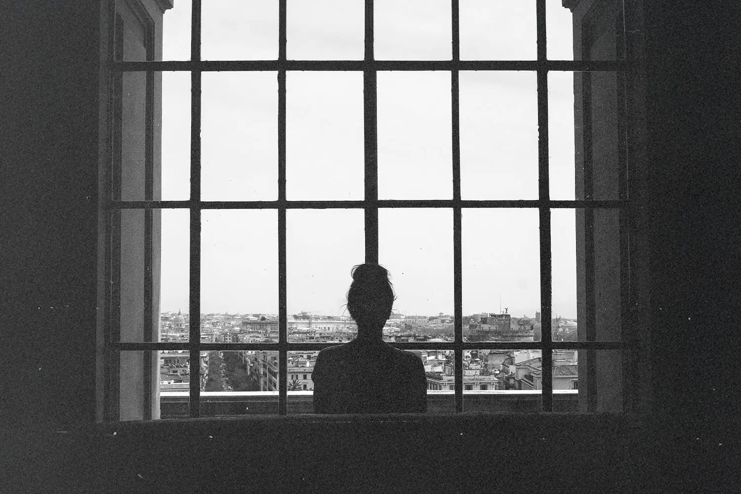 A person standing near a large window looking out over a city.