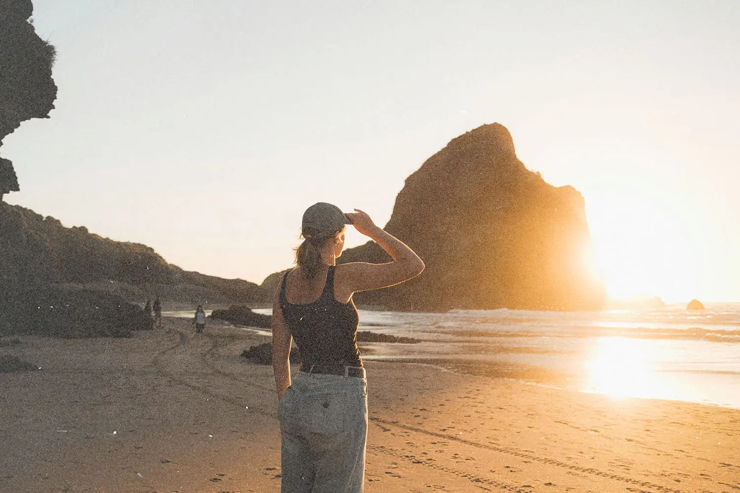 A person standing on a sandy beach facing large rock formations at sunset.