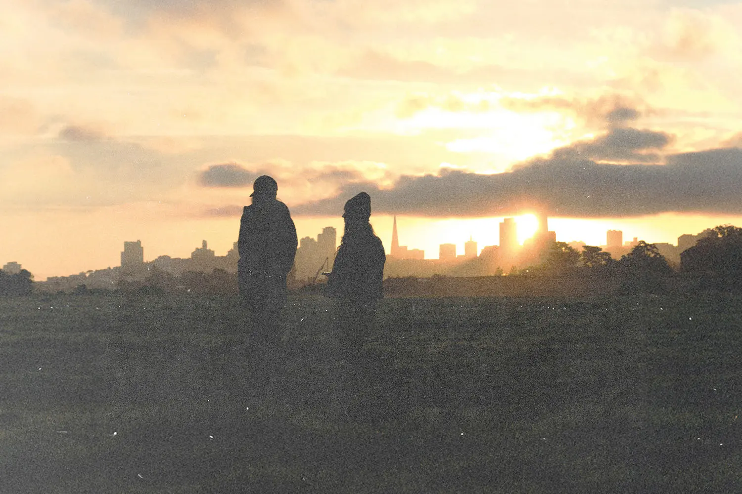 Two people standing on a hill at sunset with a city skyline in the background.