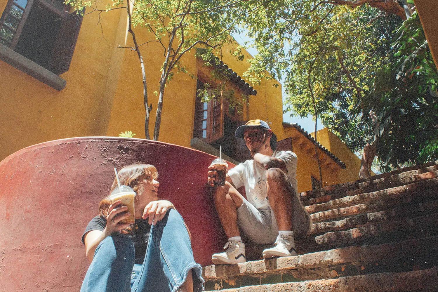 Two people sitting on outdoor steps holding drinks in a sunlit courtyard.