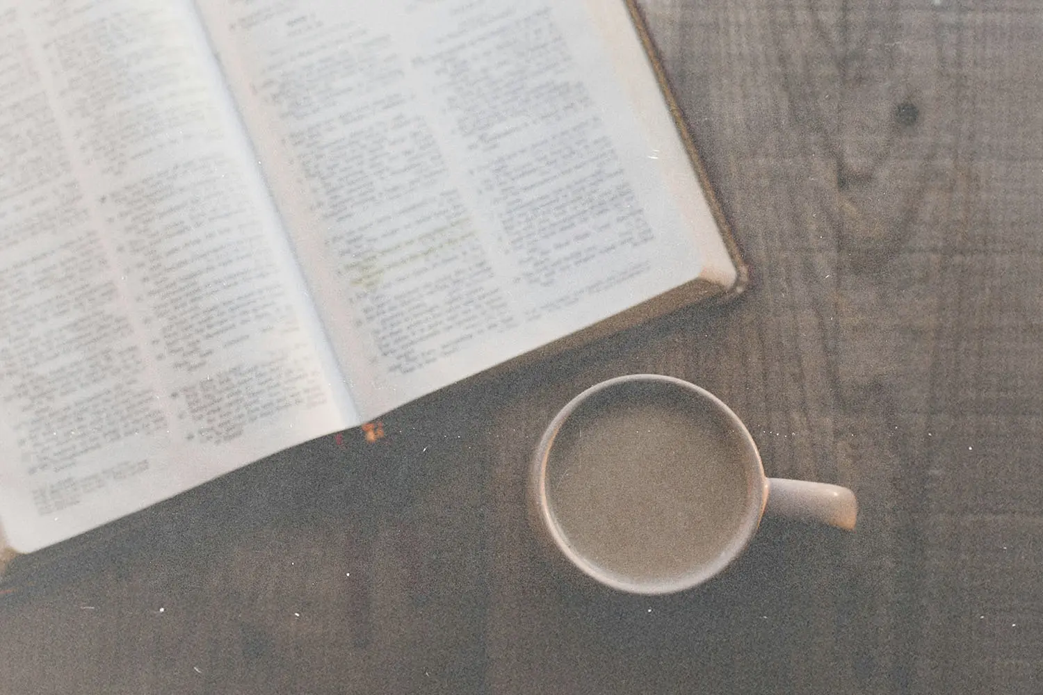 An overhead view of an open Bible beside a coffee cup on a wooden surface.