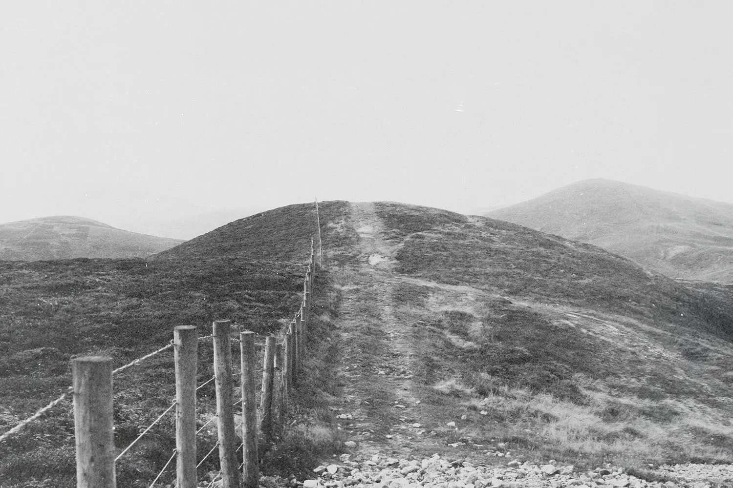 A black-and-white outdoor scene of a narrow pathway divided by fence posts across rolling hills.