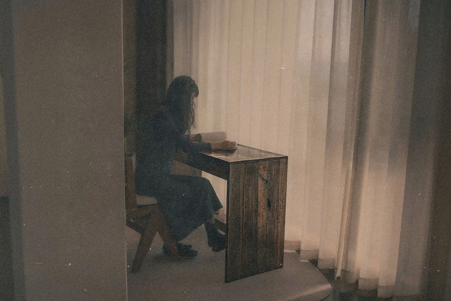 A person seated at a small wooden desk near sheer curtains with soft indoor light.