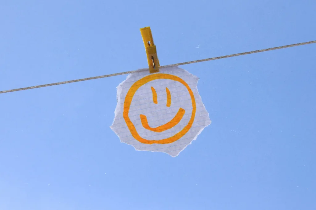 Hand‑drawn smiley face on a torn piece of paper clipped to a clothesline against a clear blue sky