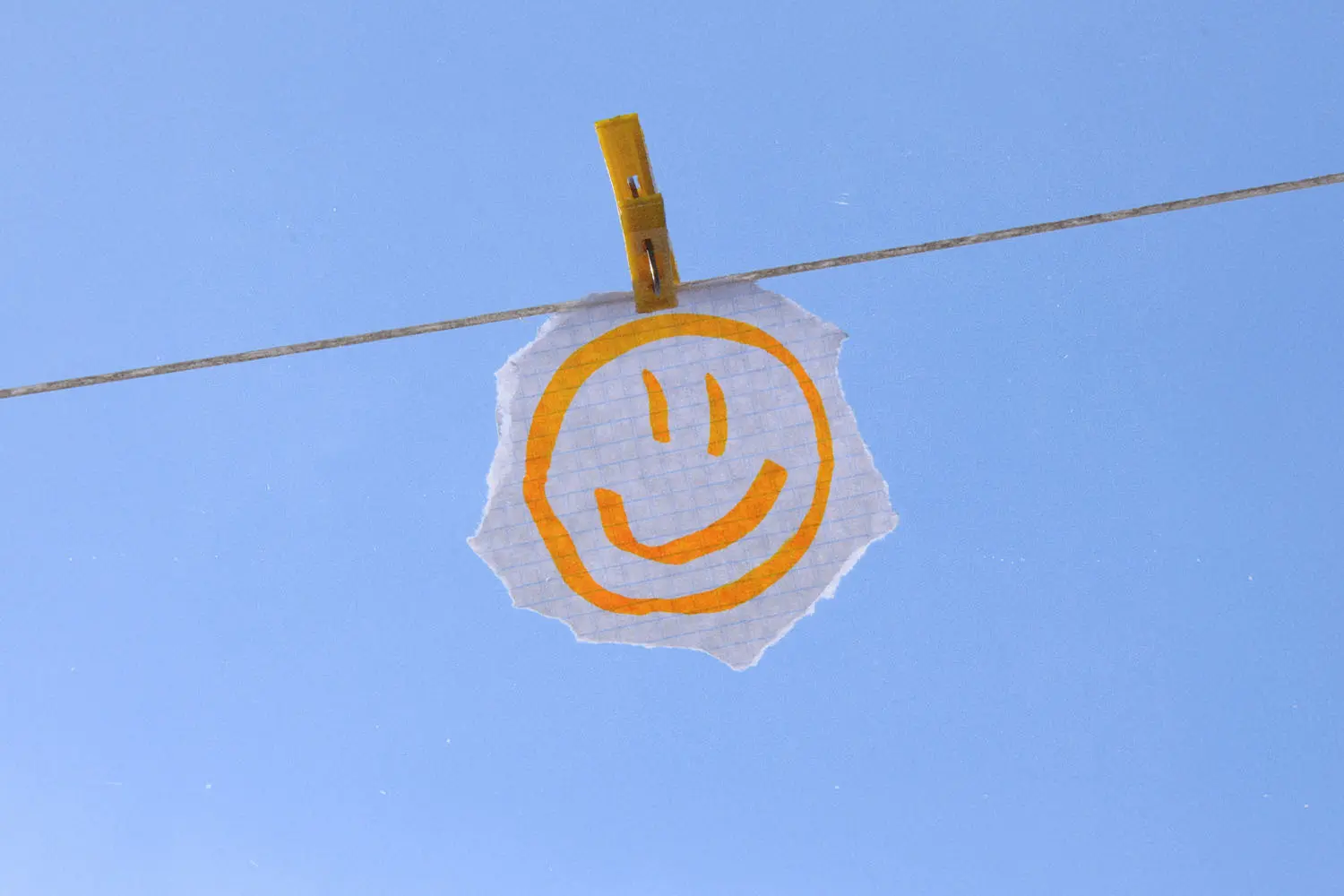 Hand‑drawn smiley face on a torn piece of paper clipped to a clothesline against a clear blue sky