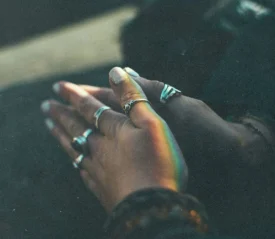 Hands clasped together in soft light, adorned with silver rings and touched by a faint rainbow reflection
