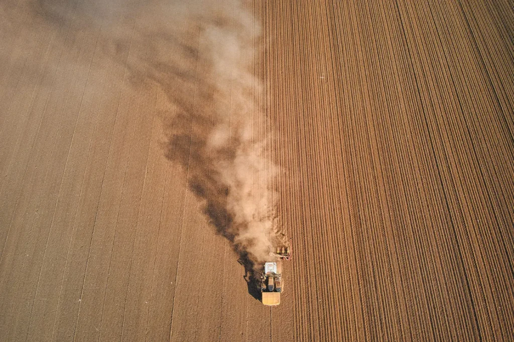 A tractor moves across a large plowed field, leaving a long trail of dust behind it