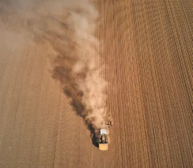 A tractor moves across a large plowed field, leaving a long trail of dust behind it