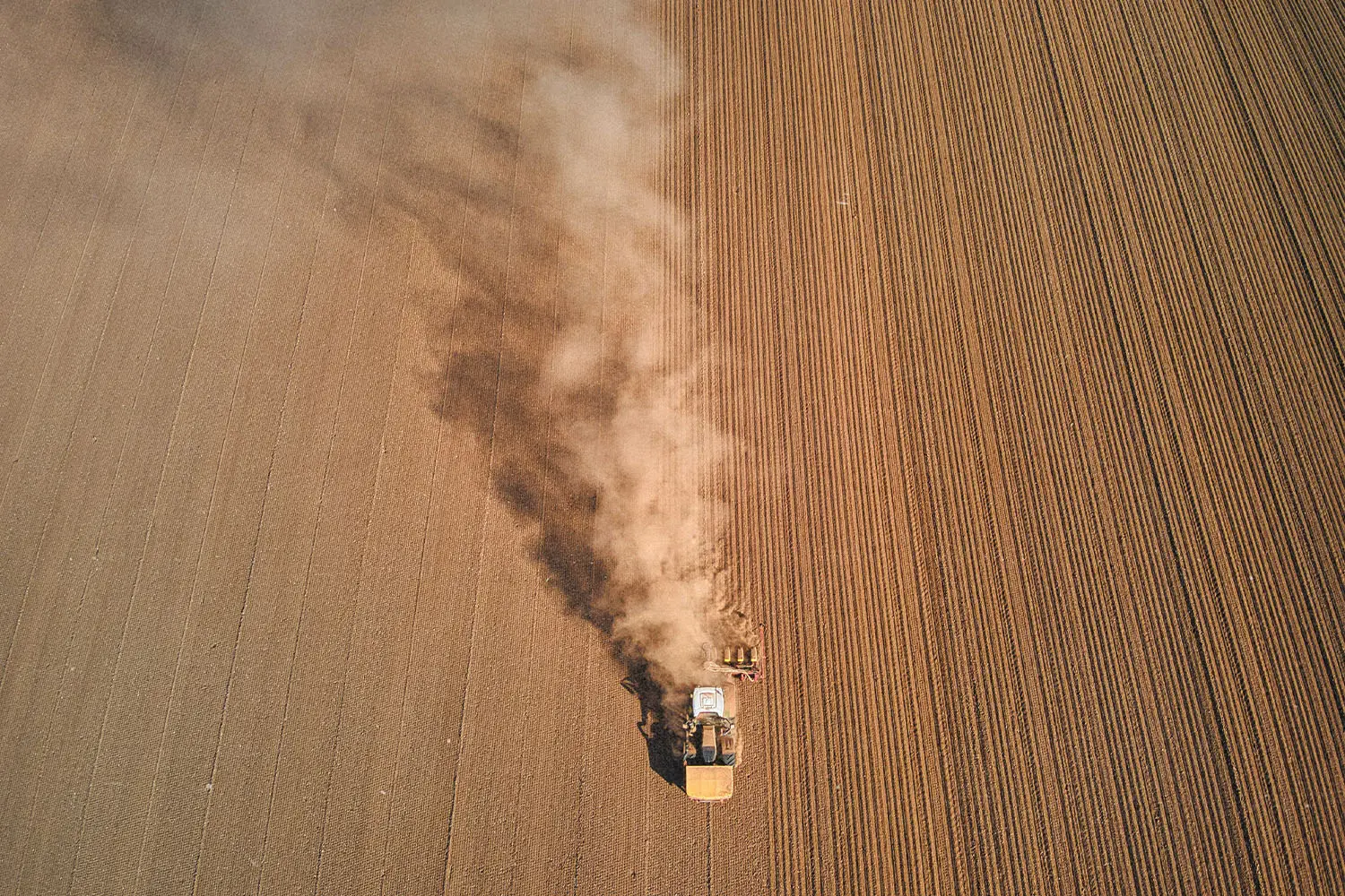 A tractor moves across a large plowed field, leaving a long trail of dust behind it