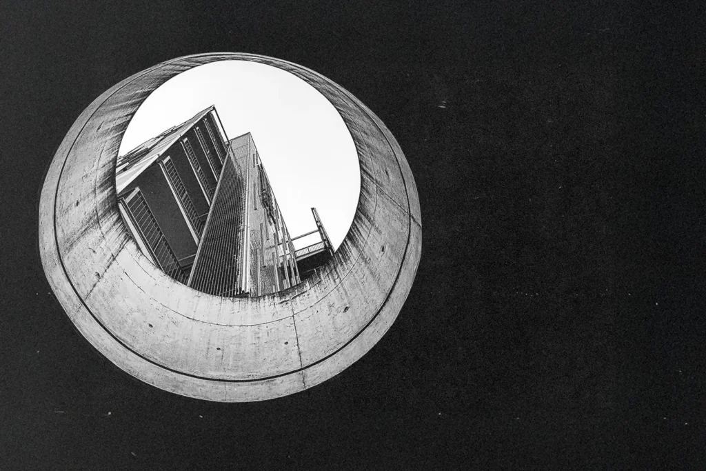 Black‑and‑white photograph looking upward through a large circular concrete opening toward a tall modern building, showing sharp architectural lines and high contrast
