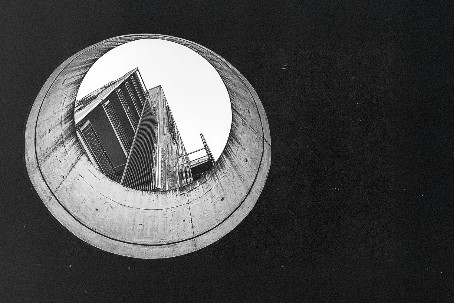 Black‑and‑white photograph looking upward through a large circular concrete opening toward a tall modern building, showing sharp architectural lines and high contrast